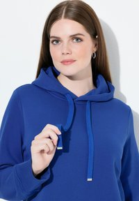 Niebieska bluza z kapturem wykonana z teksturowanej tkaniny, z prążkowanymi mankietami i sznurkami ze metalowymi zakończeniami. Model trzyma sznurek, zwrócony twarzą do przodu.