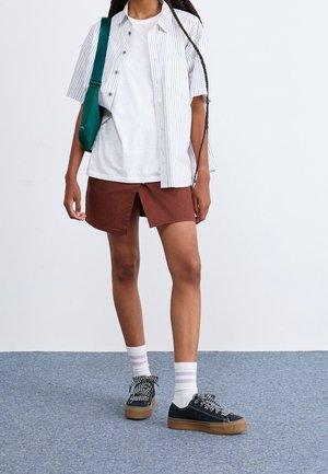 Personne portant un t-shirt blanc et une chemise à manches courtes rayée, une jupe marron avec une fente devant, des chaussettes blanches, des baskets noires et un sac bandoulière vert.
