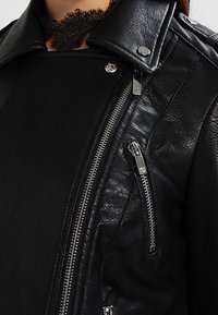 Giacca da moto in pelle nera con colletto a scasso, molteplici cerniere e finitura texture arricchita da dettagli in metallo argentato.