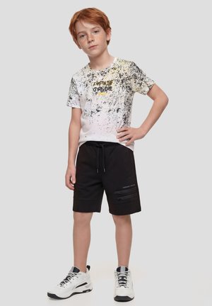 Niño pelirrojo de pie con la mano en la cadera, lleva una camiseta blanca con estampado salpicado, pantalones cortos negros con cremalleras y zapatillas blancas.