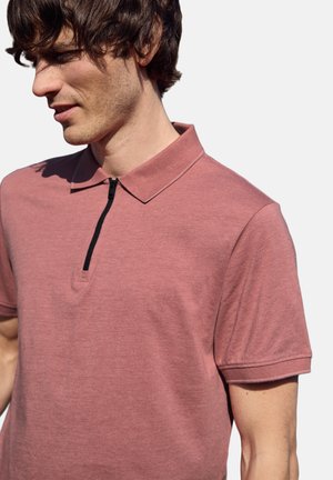 Joven con cabello oscuro que lleva una camiseta tipo polo de manga corta color rosa polvoriento con una cremallera negra sobre un fondo blanco.