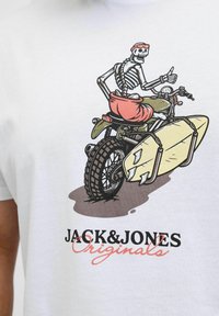 Camiseta de algodón blanca con un gráfico de un esqueleto en motocicleta con una tabla de surf, acentuada por diseños coloridos y el nombre de la marca.