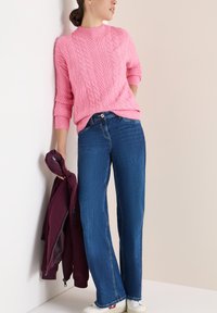 Rosa Kabelstrickpullover mit gerippten Bündchen, blaue gerade geschnittene Jeans und eine kastanienbraune Jacke in der Hand gehalten. Weiße Sneaker mit Akzenten.