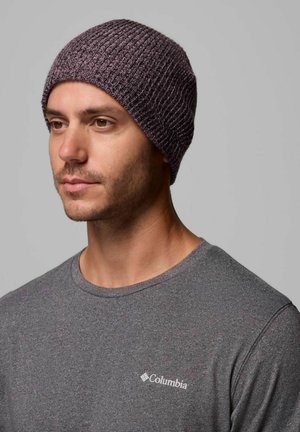 Uomo adulto che indossa un berretto viola lavorato a maglia e una t-shirt grigia Columbia, guarda leggermente a sinistra su uno sfondo grigio uniforme.