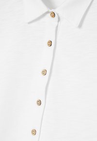 Biała koszulka polo z teksturowanej tkaniny, z klasycznym kołnierzykiem i drewnianymi guzikami z przodu.