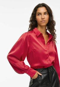 Woman with curly hair wearing a red satin blouse with puffed sleeves tucked into high-waisted black leather pants, hand in pocket.