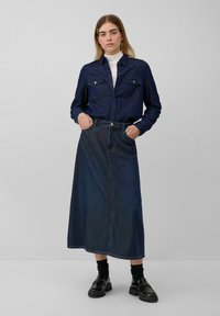 Camicia di jeans con due tasche sul petto, abbinata a una gonna lunga in denim scuro. Il modello indossa mocassini neri e calze a metà polpaccio. Sfondo neutro.