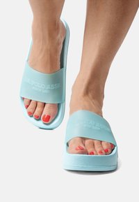 Light blue slide sandals featuring a textured rubber sole and a wide strap with embossed logo. Open-toe design reveals red-painted toenails.