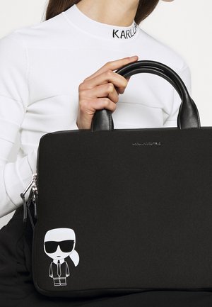 Personne portant un haut blanc à manches longues avec "KARL" sur le col, tenant un sac à main noir orné d'un personnage de dessin animé avec des lunettes de soleil et une queue de cheval.