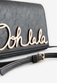 Juoda dirbtinės odos rankinė, puošta auksiniu „Ooh lala“ užrašu. Komplekte juodas dirželis su auksine sagties detalė.