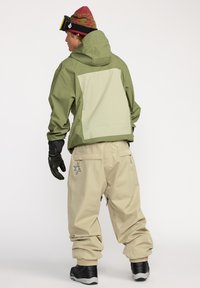 Waterdichte groene jas met een beige achterkant, gecombineerd met beige baggy broeken met ritszakken en een sterpatch, gedragen met zwarte handschoenen.