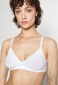 Soutien-gorge blanc sans fil avec un design triangulaire, texture lisse et fines bretelles réglables. Présente des coutures discrètes le long des bords.