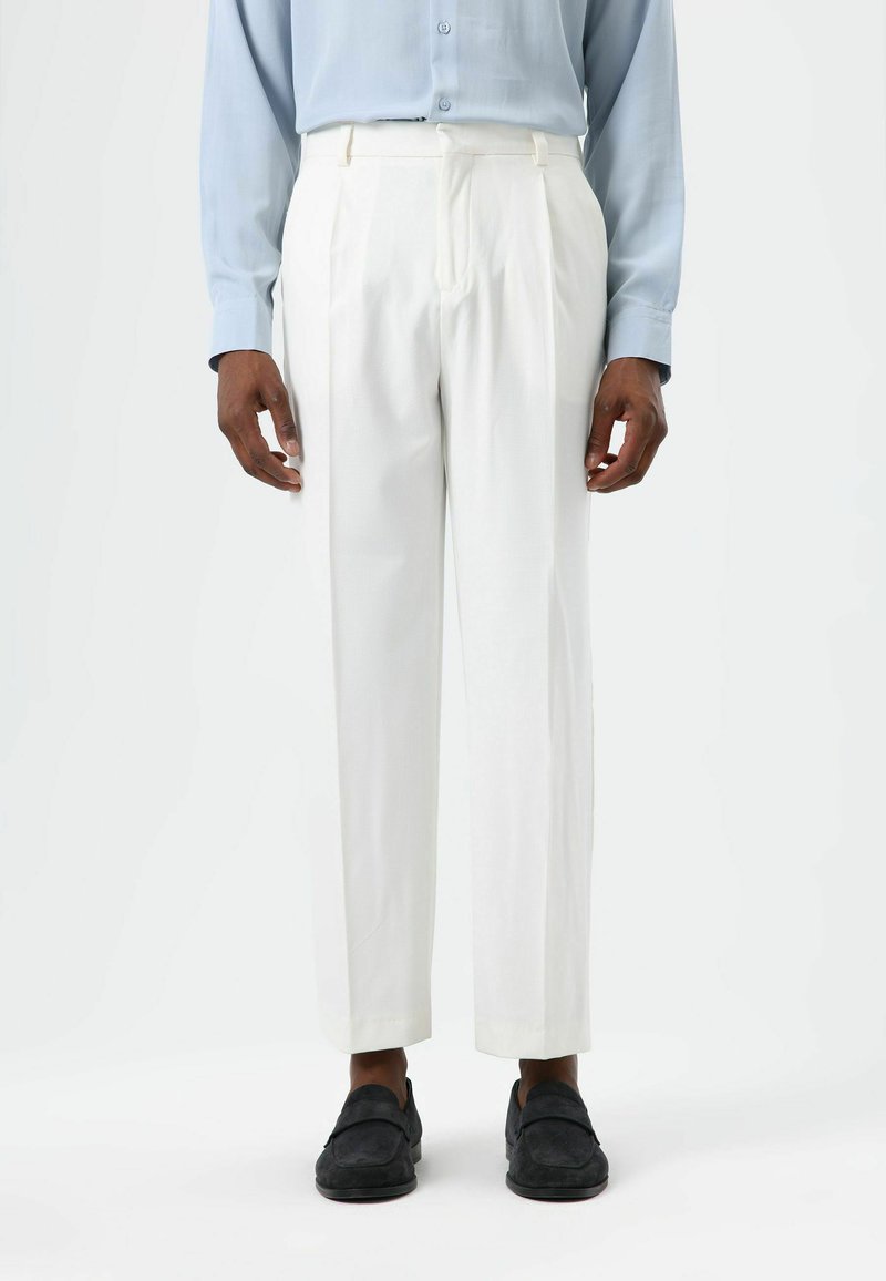 White flat-front trousers with pleats, tailored fit, and smooth texture, paired with black suede loafers. Buttons and zipper closure detail.