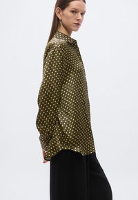 Melna satīna krekls ar zelta ģeometrisku rakstu, garām piedurknēm un vaļēju piegriezumu. Redzams izteikts spīdums un gluda tekstūra.