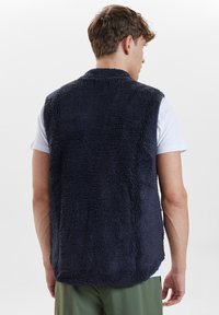 Marintröja i fleece med strukturerad yta, ärmlös design och rundad nederkant. Bärs över en vit skjorta och har en avslappnad passform.