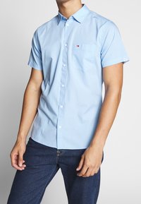 Ljusblå kortärmad skjorta, knäppning framtill, ficka på vänster bröst, har logotypetikett på ärmen, tillverkad av mjuk bomullstyg.