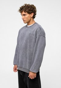 Grijze sweatshirt van zacht materiaal, met een ronde hals, verlaagde schouders en subtiele logodetail op de borst. Zwarte losse broek.
