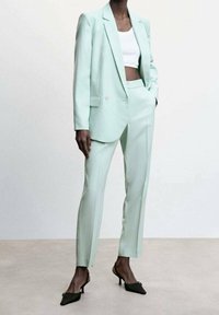 Mintgroene maatpak met een dubbelzijdige blazer en slim-fit broek. Soepele textuur, gecombineerd met een witte cropped top en zwarte hakken.