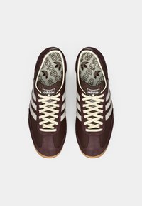 Sneakers in suede e mesh marrone con strisce e lacci bianchi. Logo in evidenza sulla linguetta e fodera interna a fantasia. Suola in gomma.