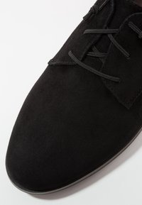 Chaussure en daim noir avec une texture lisse, présentant un design classique à lacets et un bout rond. La semelle extérieure est sombre, complétant la tige.