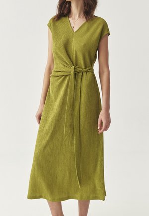 Femme portant une robe midi verte texturée sans manches, avec un col en V et une ceinture nouée à la taille, sur un fond uni.
