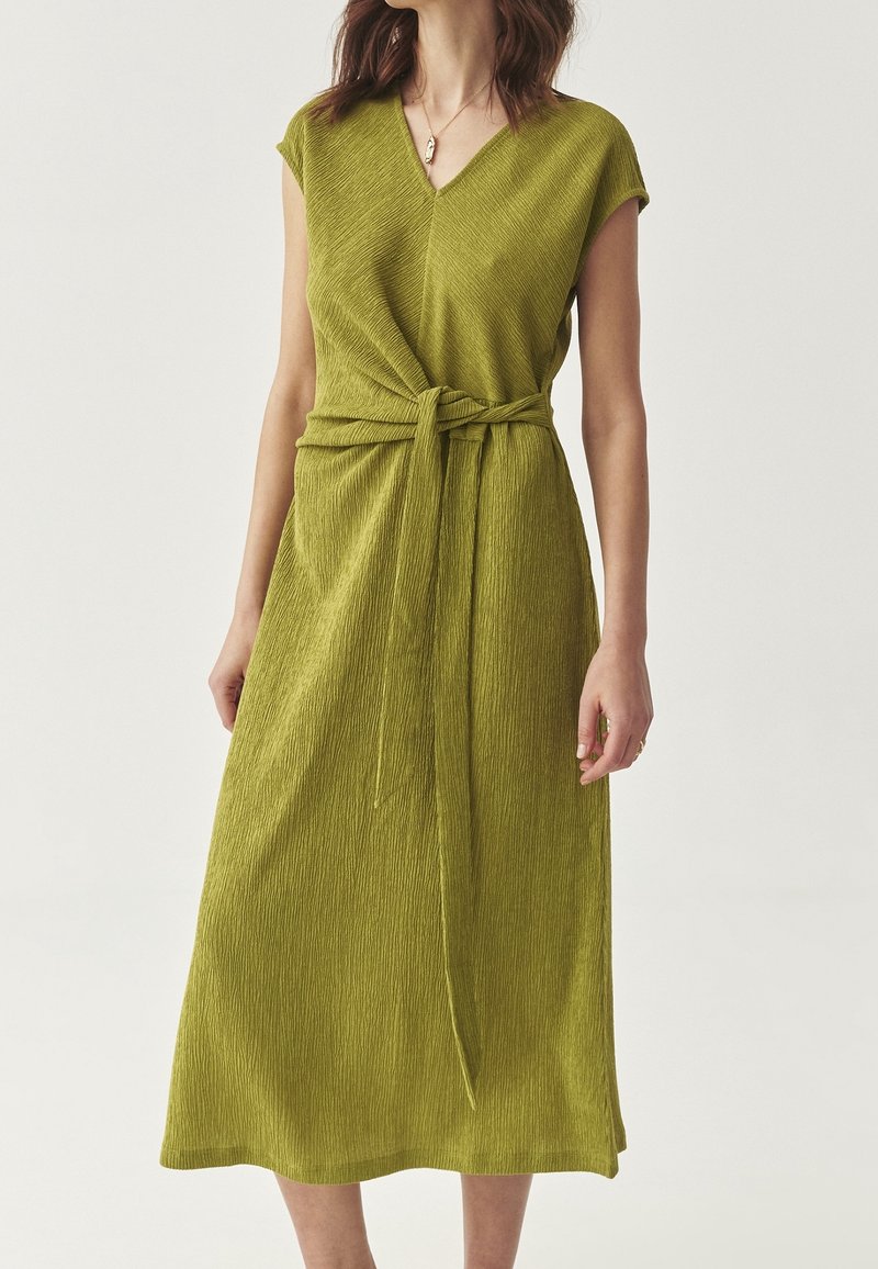 Femme portant une robe midi verte texturée sans manches, avec un col en V et une ceinture nouée à la taille, sur un fond uni.
