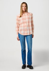 Camicia a maniche lunghe con bottoni, a motivo check in arancione e crema, indossata con pantaloni flare in denim blu e zoccoli neri.