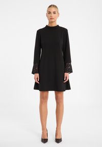 Schwarzes Kleid mit hohem Kragen und schwingenden Ärmeln, die mit Spitzenakzenten versehen sind. Hergestellt aus glattem Stoff, hat es ein schwingendes Rockdesign.