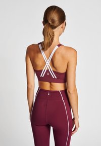 Bordowy stanik sportowy z białymi krzyżującymi się ramiączkami oraz dopasowane legginsy o wysokim stanie, z białymi paskami po bokach i gładką teksturą.