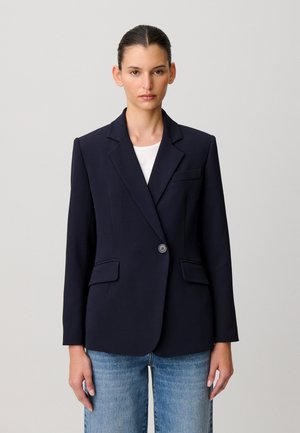 Mujer joven con el cabello peinado hacia atrás, lleva una blazer azul marino de un solo botón sobre una camisa blanca y jeans azules, de pie frente a un fondo liso.