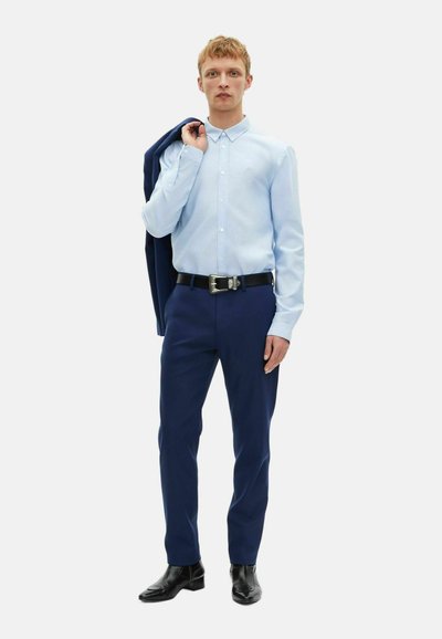 Helles blaues Hemd mit Knöpfen, dunkelblaue Hose, schwarzer Gürtel mit silberner Schnalle, schwarze Schuhe, navyfarbene Jacke über der Schulter haltend, neutraler Hintergrund.