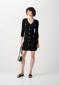 Robe noire en tricot avec un décolleté en V, des manches trois-quarts, des boutons sur le devant, deux poches et une silhouette ajustée. Assortie avec des sandales à talons blocs noires.