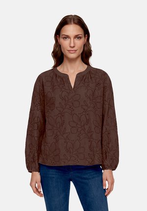 Femme portant un chemisier marron à manches longues avec une broderie florale et un petit col en V, associée à un jean bleu, debout devant un fond uni.