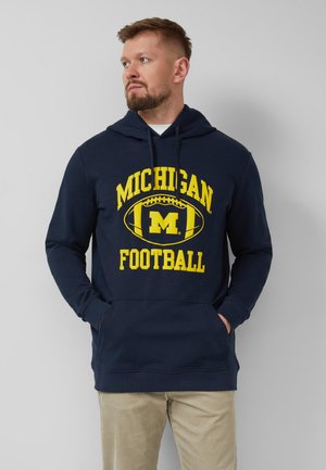 Mężczyzna z brodą w granatowej bluzie z kapturem Michigan Football i beżowych spodniach, ręce w kieszeniach, patrzący w lewo na jasnym, jednolitym tle.