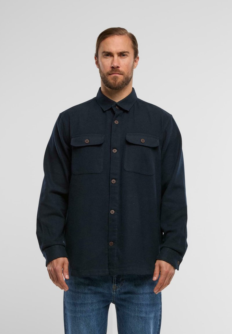 Sötét tengerészkék, textúrált anyagból készült gombos ing, két mellzsebbel és barna gombokkal. Szabványos gallér és hosszú ujjú.