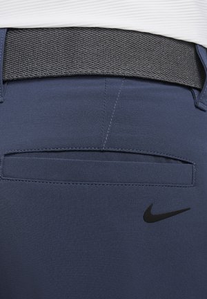 Tamsiai mėlynos sportinės kelnės su tekstūruotu pilku juosmeniu, turinčios užpakalinę kišenę ir juodą Nike logotipą apatinėje pusėje.