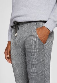 Calças cinzentas estampadas com cintura ajustável e bolso lateral. O tecido apresenta um design de xadrez texturizado e um brilho subtil.