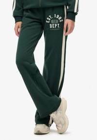 Groene katoenen sweatpants met een uitlopende pasvorm, crèmekleurige zijstrepen en een bedrukte logo met de tekst "EST. 1982 SD DEPT. SUPERDRY." Accenten zijn minimaal.