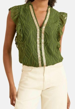 Blouse verte sans manches avec motif de feuilles texturé, épaules volantées et bordure en sequins dorés sur le devant, portée avec un pantalon écru.