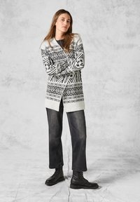 Strikket cardigan med et geometrisk svart-hvitt mønster, åpen front og ribbestrikkede manchetter, parret med mørke jeans og svarte snøresko.