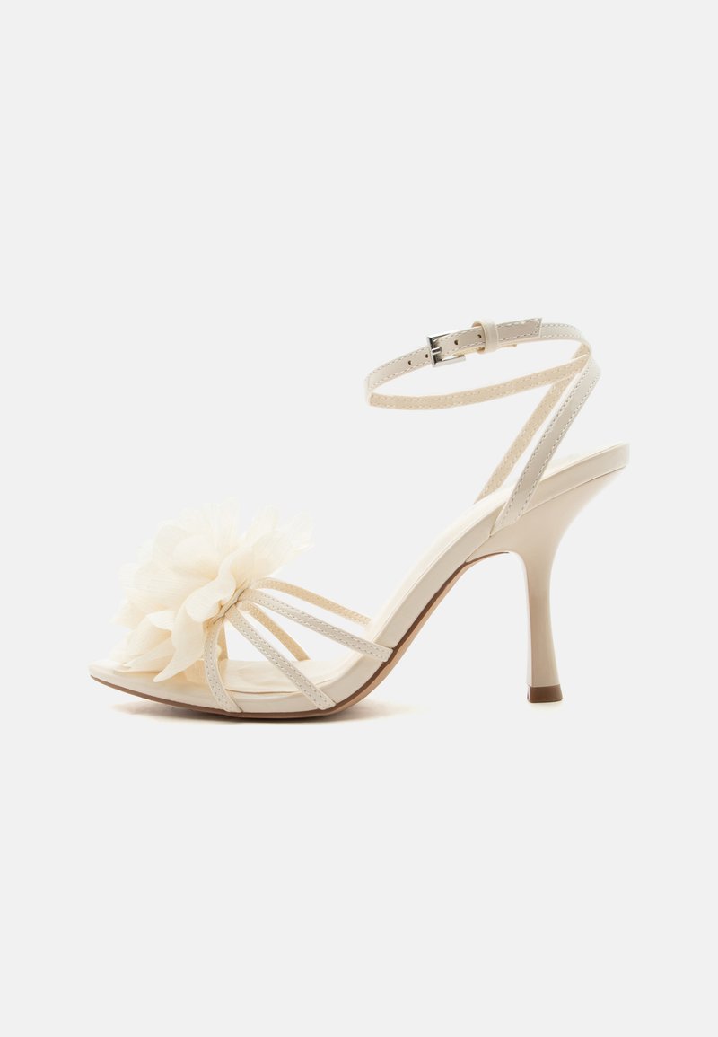 Λευκό πέδιλο με ψηλό τακούνι, λεπτό λουράκι στον αστράγαλο, ανοιχτό δάχτυλο και μεγάλη υφασμάτινη λουλουδένια λεπτομέρεια στις μπροστινές λουρίδες.