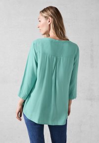 Türkise Bluse mit Dreiviertelärmeln, rundem Ausschnitt, plissierter Rückendesign und glattem, fließendem Stoff. Kombiniert mit Jeans.