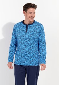 Langärmliges Hemd in Blau mit einem Blattmuster, ausgestattet mit einem Rundkragen und einer Knopfleiste mit zwei Knöpfen. Kombiniert mit marineblauen Hosen.