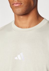 Hellgraues T-Shirt mit Rundhalsausschnitt, das links auf der Brust ein kleines weißes gesticktes Logo mit drei Streifen trägt.