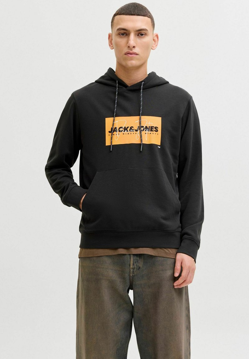 Jeune homme aux cheveux courts portant un sweat à capuche noir avec le logo Jack & Jones orange et un jean marron délavé, debout devant un fond blanc.