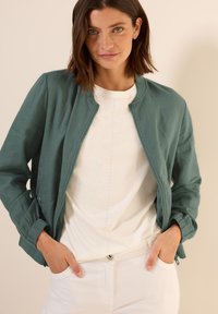 Turquoise bomberjack met elastische cuffs, lichtgewicht stof, ritsluiting, gedragen over een witte blouse met decoratieve studs, en gecombineerd met witte broek.