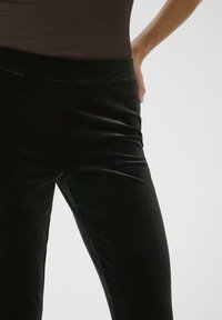 Pantalons en velours noir avec une texture lisse et une silhouette ajustée ; présentent une taille haute et ne comportent ni fermetures ni poches visibles.