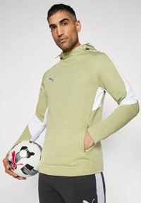 Lichtgroene hoodie met witte accenten, voorzien van een kangoeroezak en Puma-logO. Model houdt een zwart-witte voetbal vast.