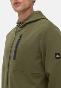 Sweatshirt à capuche vert olive avec une poche poitrine zippée, un patch logo noir et orange contrastant, et un tissu doux et texturé.