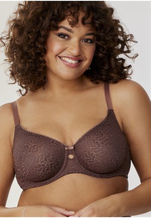Smiling woman with curly hair wearing a brown lace bra, with hands gently clasped below the chest against a plain background.